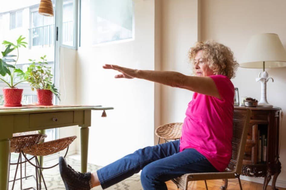 Elderly woman smiling while performing a seated arm stretch in a bright, cozy living room.