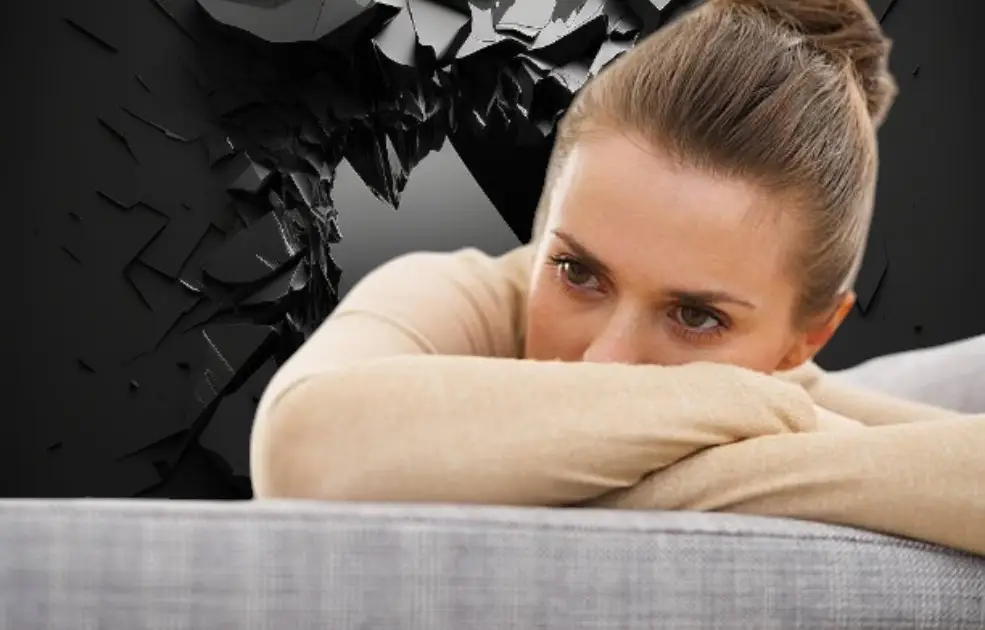 A person sits slouched on a couch in a dimly lit room, surrounded by clutter, staring blankly ahead—conveying fatigue and emotional heaviness.