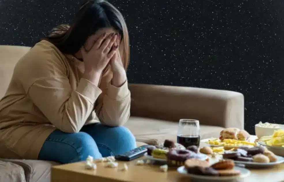 A person sits alone in a dimly lit room surrounded by junk food, with a distant, hollow expression.