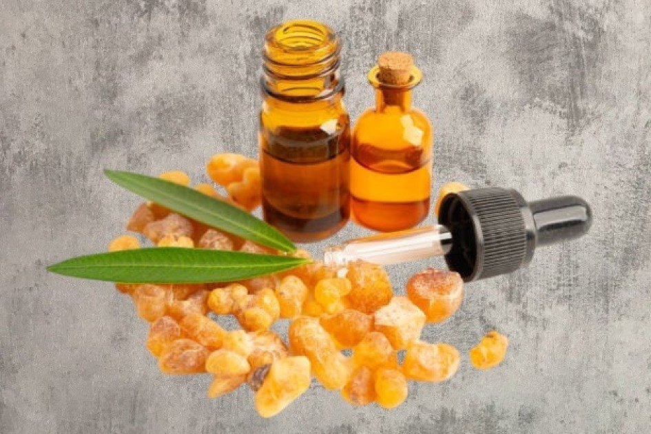 Amber bottle of frankincense essential oil beside resin and green leaves on a wooden surface.