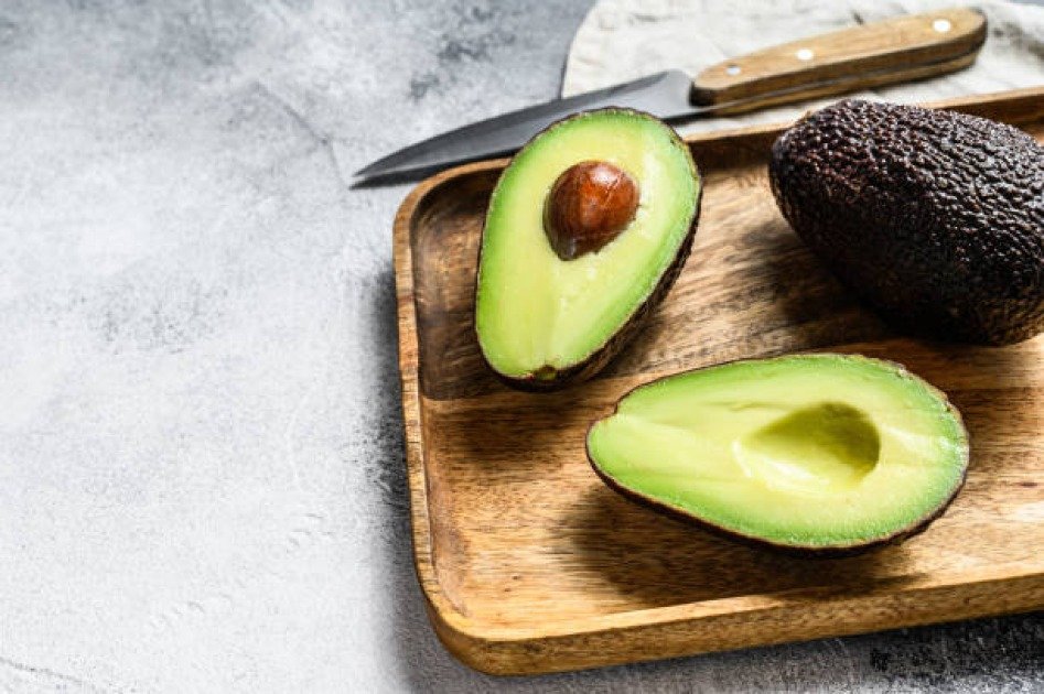 Halved avocado on a wooden board with clock hands overlaid, symbolizing intermittent fasting and healthy fats.