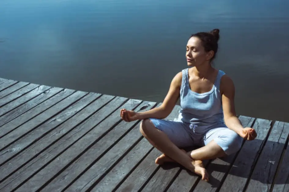 Person meditating by a tranquil lake at sunrise, surrounded by nature’s stillness.
