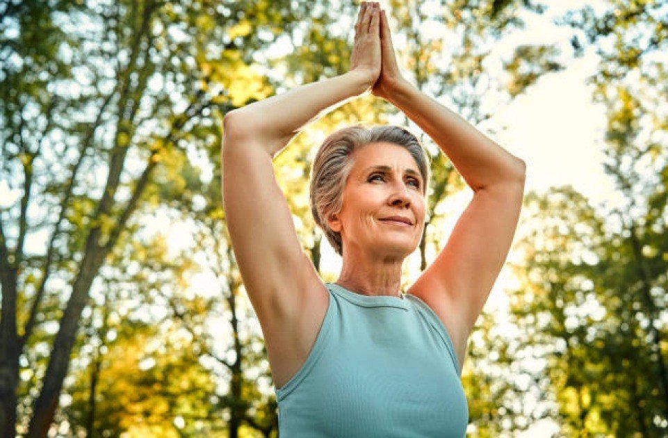 Elderly woman practicing yoga outdoors, smiling peacefully with arms raised, symbolizing wellness and mental clarity.