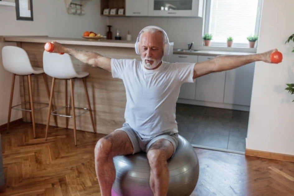 Elderly man lifting dumbbells in a bright kitchen, surrounded by fresh fruits and vegetables.