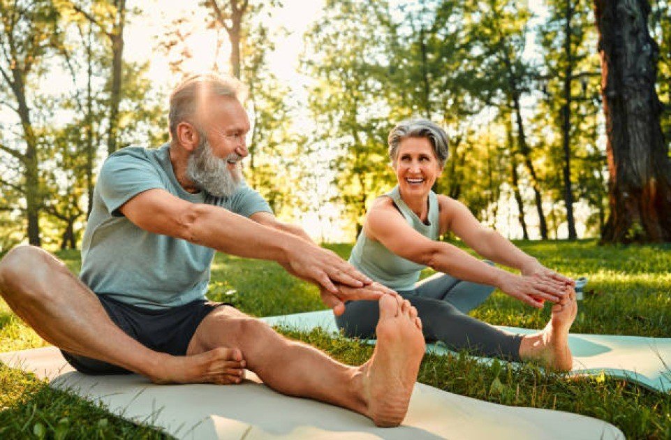 Elderly man and woman exercising together outdoors, smiling and stretching in activewear.