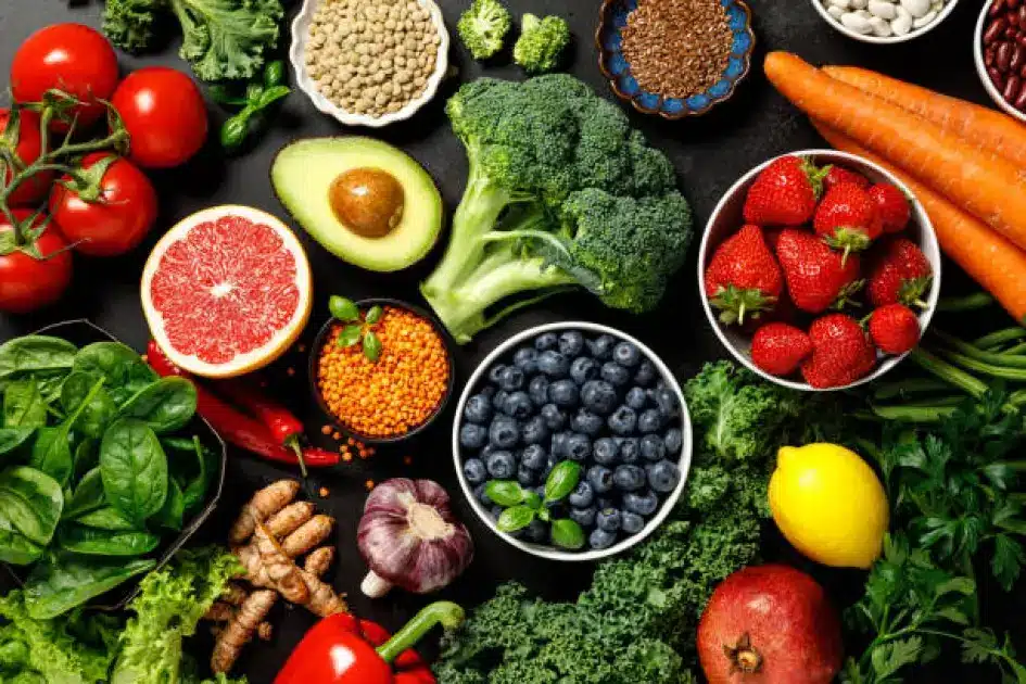 A vibrant assortment of fresh plant-based foods including leafy greens, colorful fruits, grains, and legumes arranged on a rustic wooden table.
